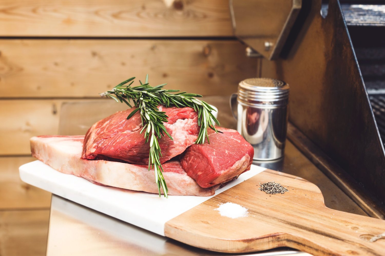 Fresh cuts of beef with rosemary garnish on a cutting board, alongside salt and pepper, in a rustic kitchen setting. Perfect for grilling or roasting.