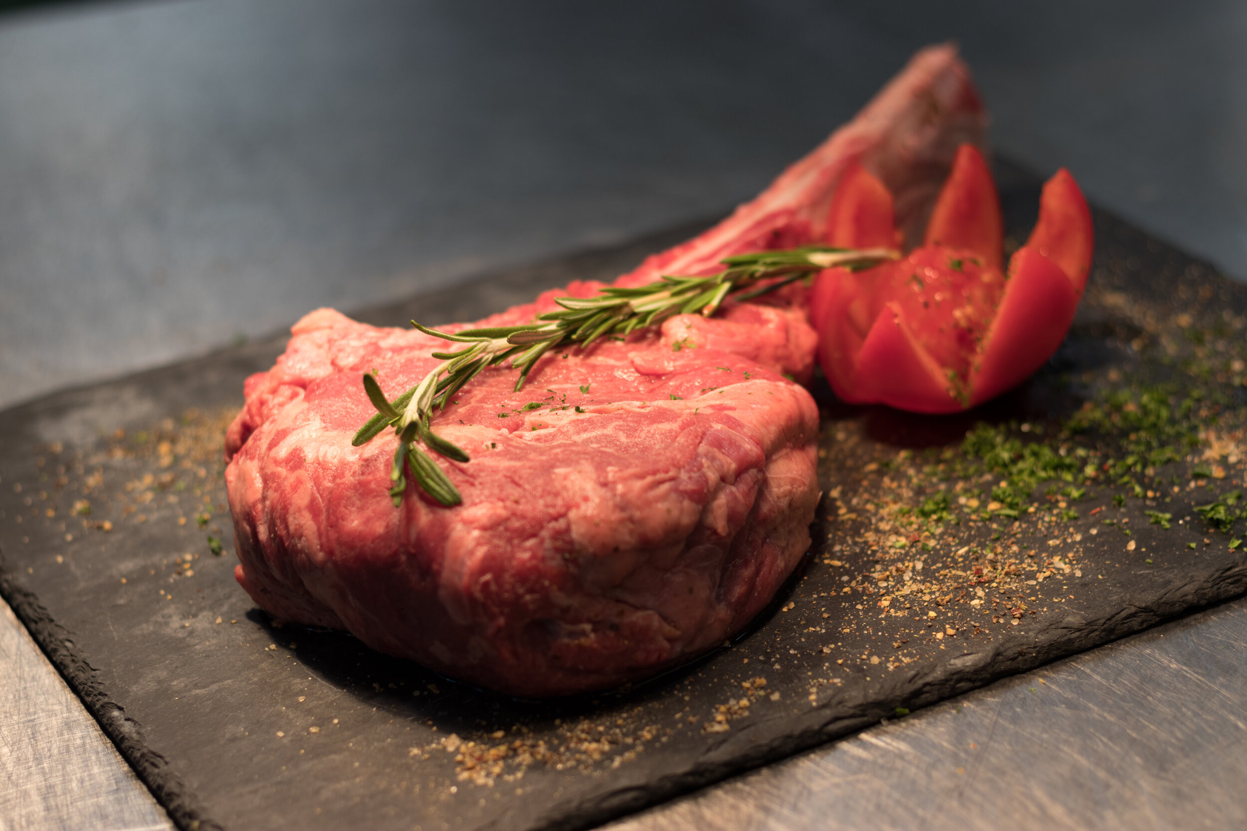 Raw beef steak with rosemary garnish, accompanied by sliced tomato, presented on a black slate surface. Ideal for culinary and food photography.