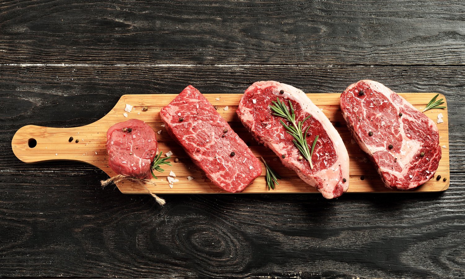Assorted cuts of raw beef displayed on a wooden cutting board, garnished with fresh rosemary and sprinkled with coarse salt and black pepper, set against a rustic black wooden background.